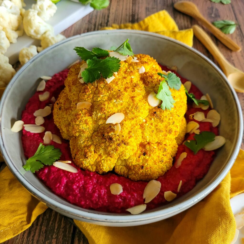 photo culinaire d'un chou-fleur entier rôti au épices avec un houmous de betterave, des amandes effilées et du citron vert